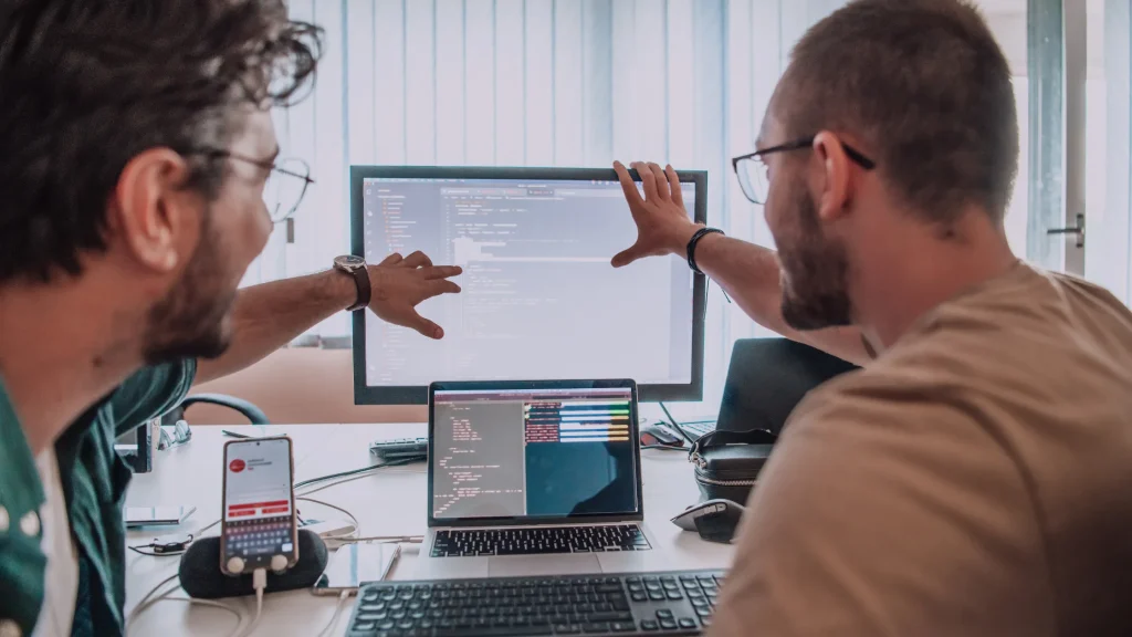 Two people collaborating, reviewing code on a computer screen, and discussing web development techniques.